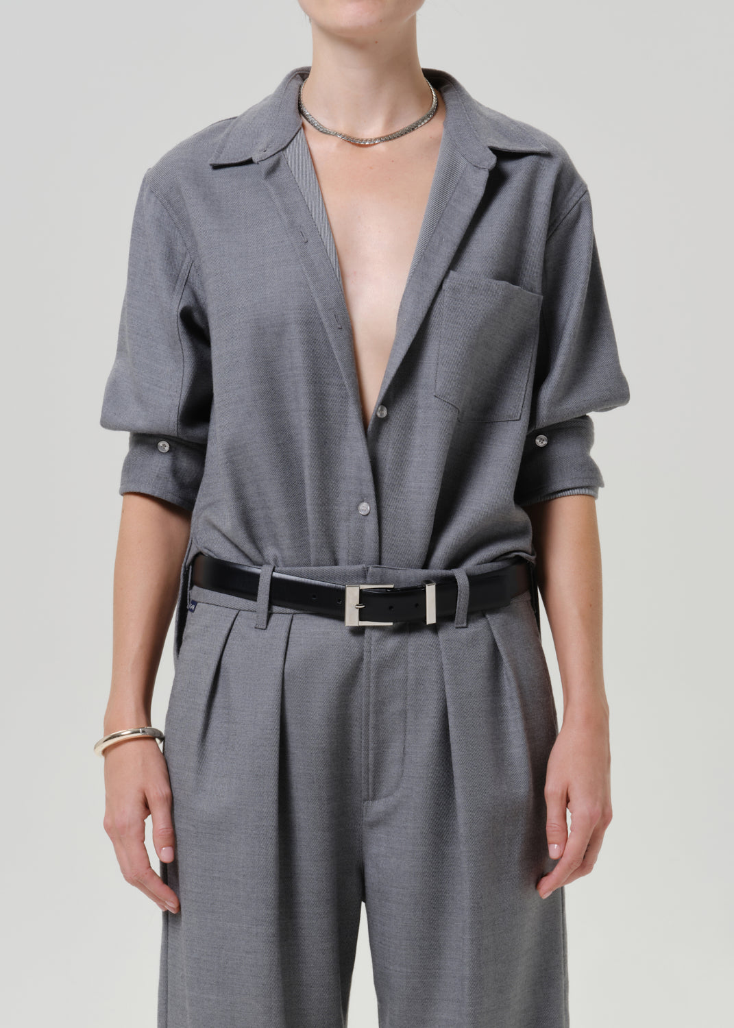 A model wearing a gray two-piece set consisting of a short-sleeve button-up shirt and matching pleated trousers, styled with a black belt and silver hardware.