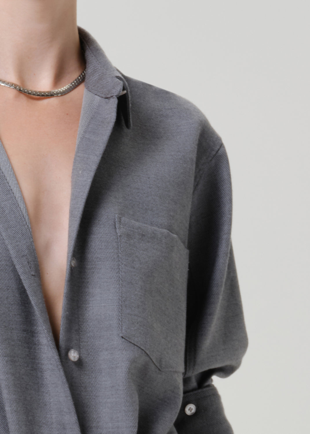 Close-up of a person wearing a gray knit button-front shirt with a collar and chest pocket, paired with a silver chain necklace, against a light gray background.