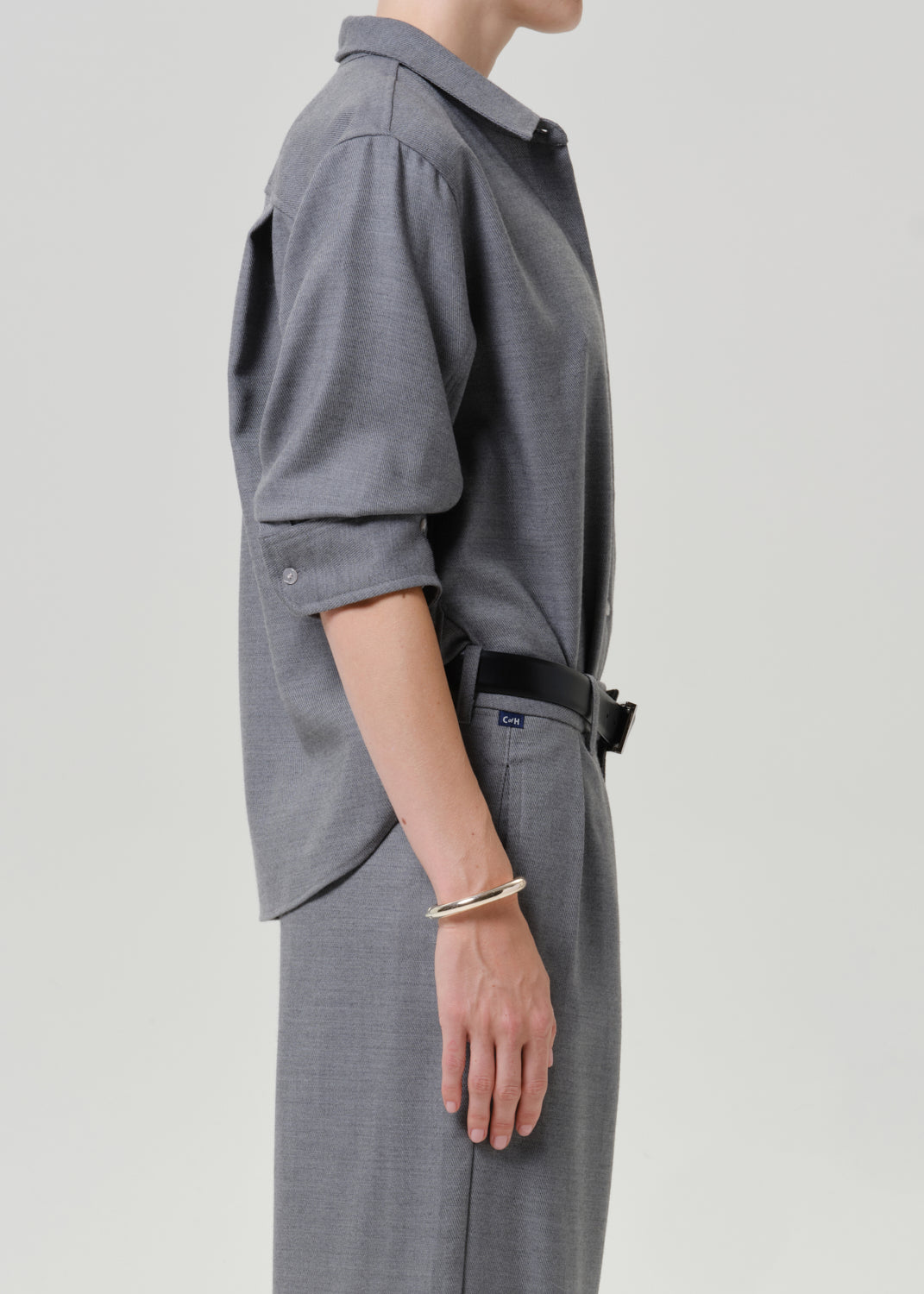 A side-profile view of a person wearing a gray long-sleeve button-up shirt with rolled sleeves and matching gray trousers, accessorized with a black belt and a gold bangle, against a light gray studio background.