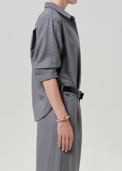 A side-profile view of a person wearing a gray long-sleeve button-up shirt with rolled sleeves and matching gray trousers, accessorized with a black belt and a gold bangle, against a light gray studio background.