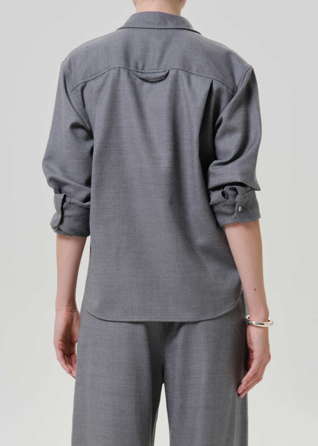 A person shown from the back wearing a gray long-sleeve button-up shirt with rolled cuffs and matching gray trousers, photographed against a plain light gray studio background.