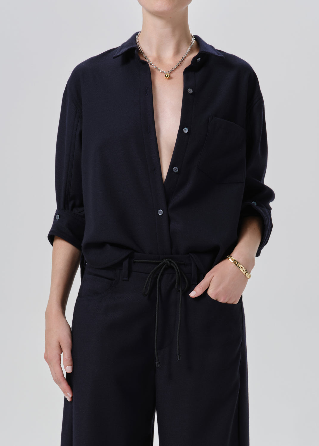 A model wearing a dark navy blue button-down shirt with rolled-up sleeves and matching dark trousers, styled with gold jewelry against a grey background.