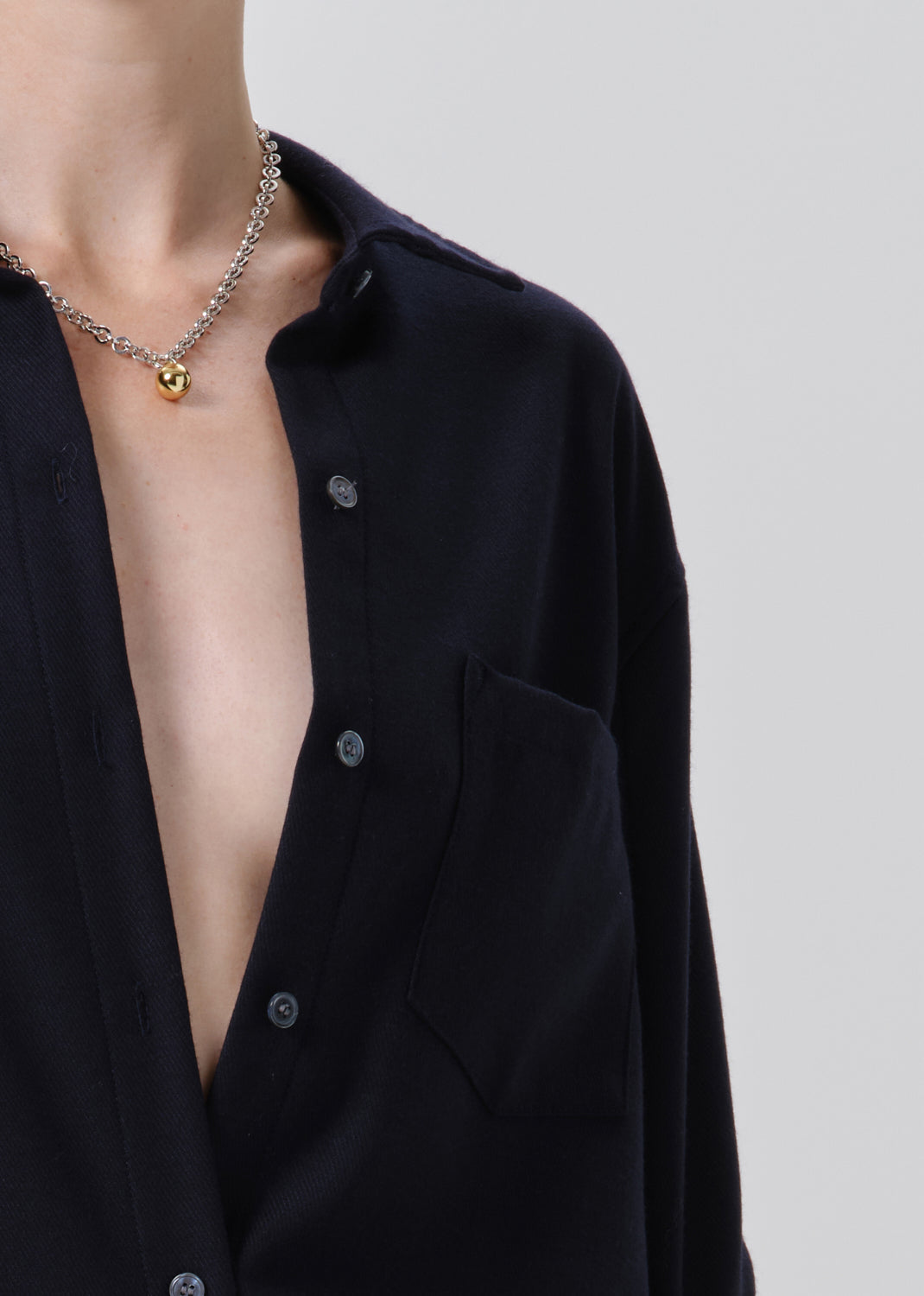 Close-up of a person wearing a black button-up shirt and a silver chain necklace with a small gold heart pendant, against a grey background.