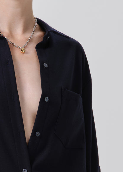 Close-up of a person wearing a black button-up shirt and a silver chain necklace with a small gold heart pendant, against a grey background.