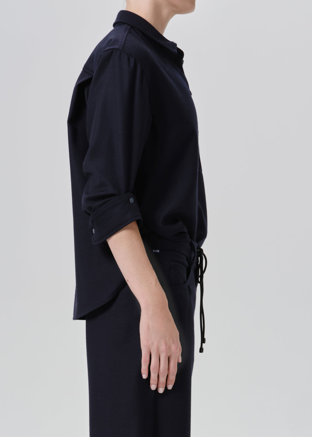 A side-profile view of a model wearing a dark navy blue shirt with rolled-up sleeves and matching dark trousers against a grey background.
