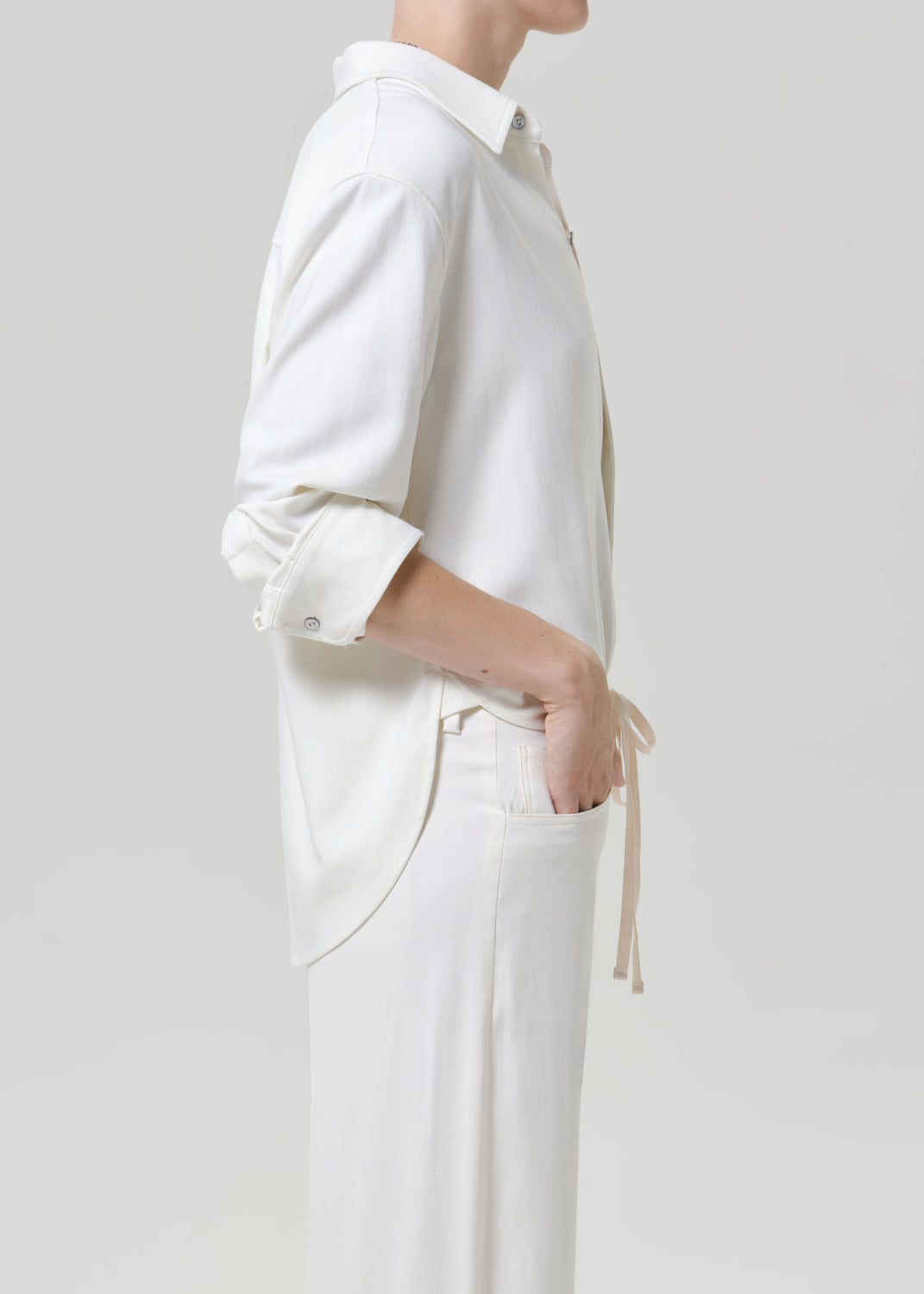 Woman wearing a white button-up shirt with white drawstring pants viewed from the side on a grey background.
