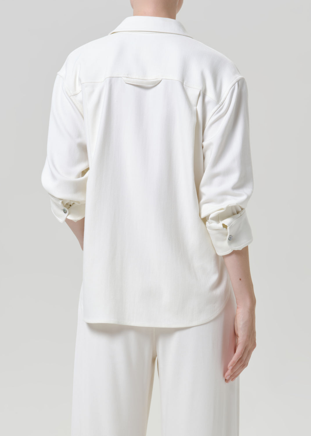 Woman wearing a white button-up shirt with white drawstring pants viewed from the back on a grey background.