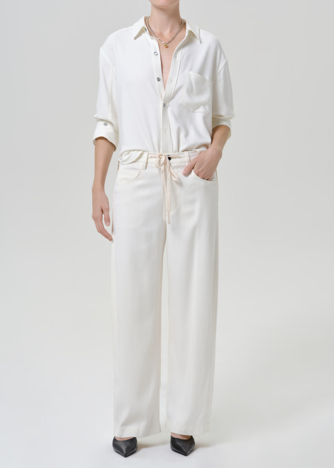 Woman wearing a white button-up shirt with white drawstring pants on a grey background.