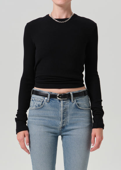 A model wearing a black long-sleeve ribbed knit crop top and light blue jeans with a black belt, shown from the neck down against a grey background.