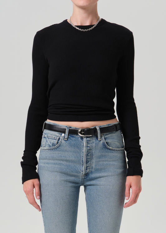 A model wearing a black long-sleeve ribbed knit crop top and light blue jeans with a black belt, shown from the neck down against a grey background.