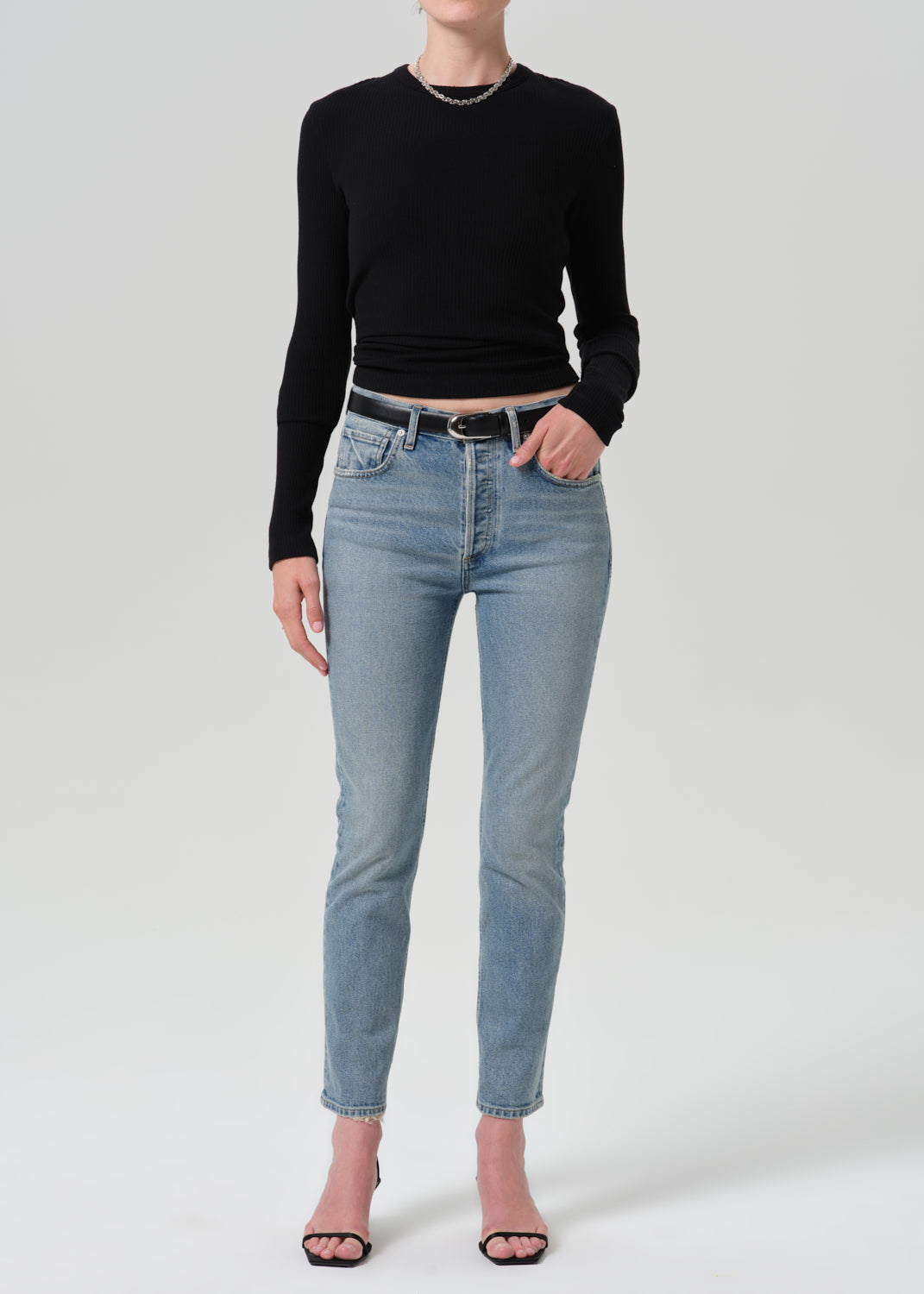 A model wearing a black long-sleeve crewneck top, light-wash blue skinny jeans with a black belt, and pink heeled sandals, standing against a grey background.