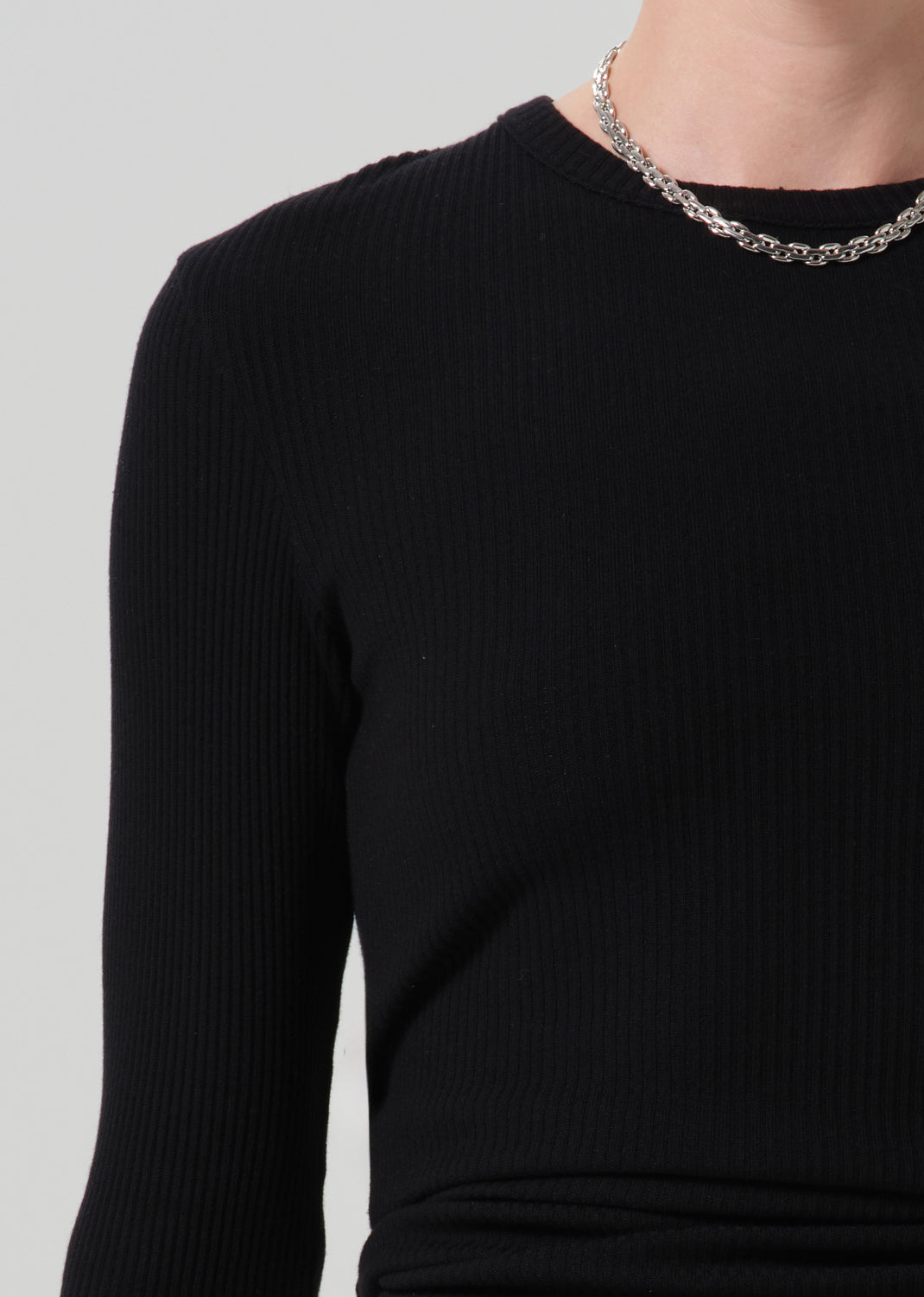 Close-up of a person wearing a black ribbed long-sleeve top and a silver chain necklace against a grey background.