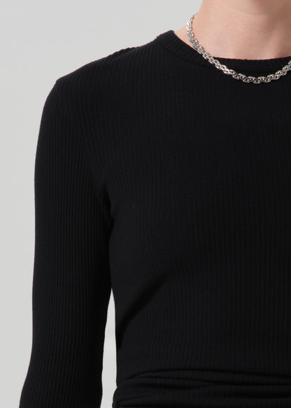 Close-up of a person wearing a black ribbed long-sleeve top and a silver chain necklace against a grey background.