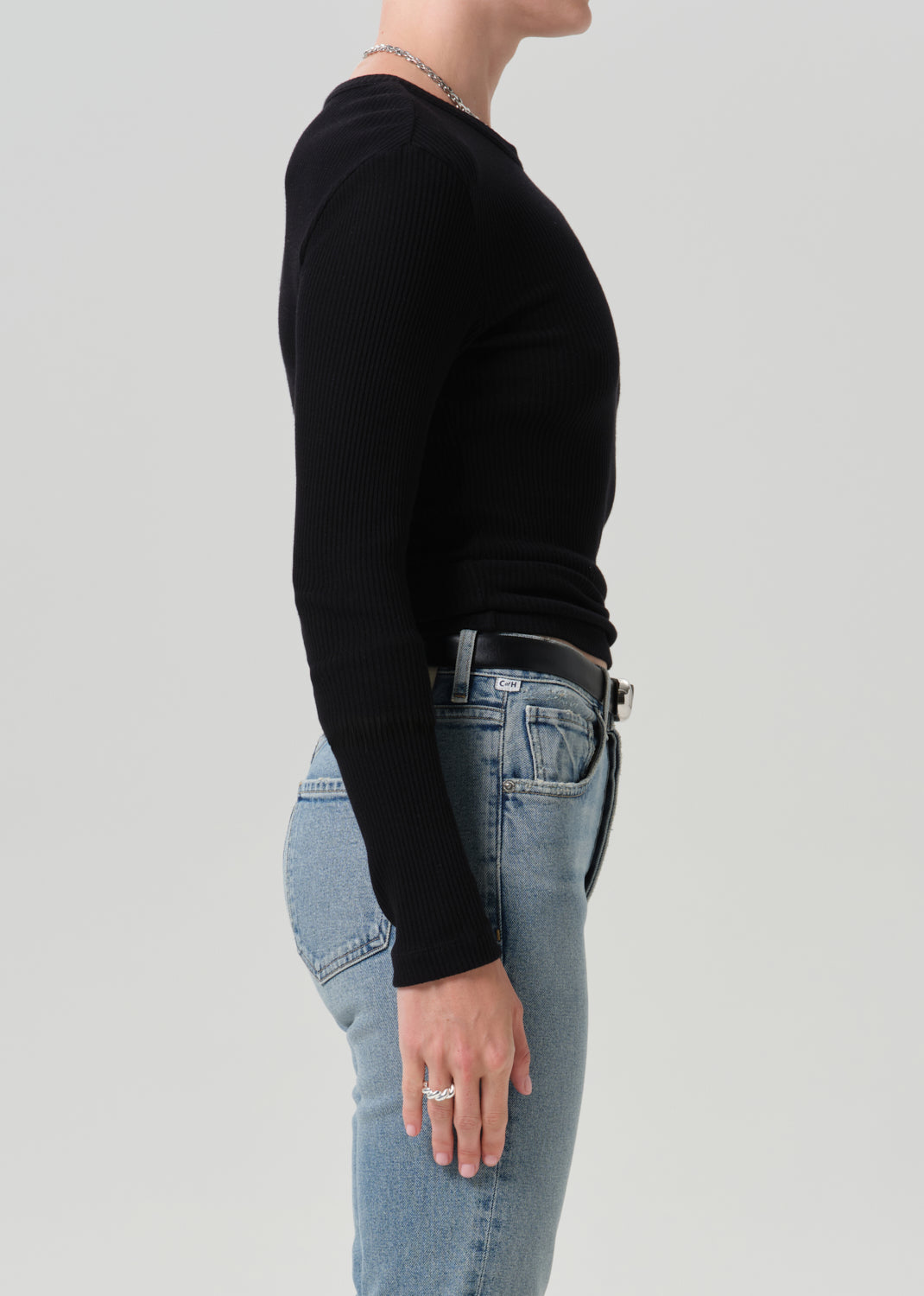 A side-profile view of a person wearing a fitted black long-sleeve ribbed top and light blue jeans against a grey background.