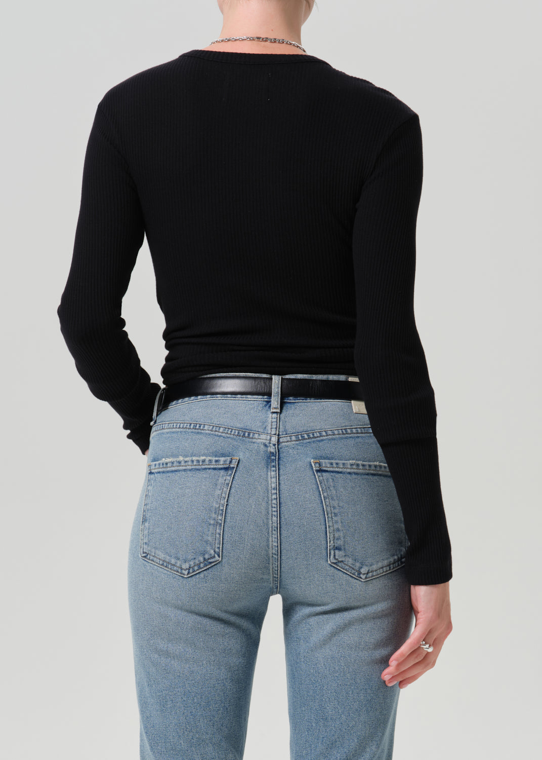 Rear view of a model wearing a black ribbed long-sleeve top and light blue jeans with a black belt, against a grey background.