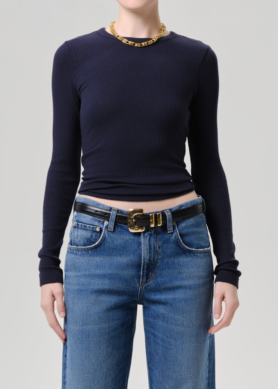 A model wearing a dark navy blue ribbed long-sleeve top tucked into blue jeans with a black belt featuring a gold buckle, accessorized with a gold chain necklace.