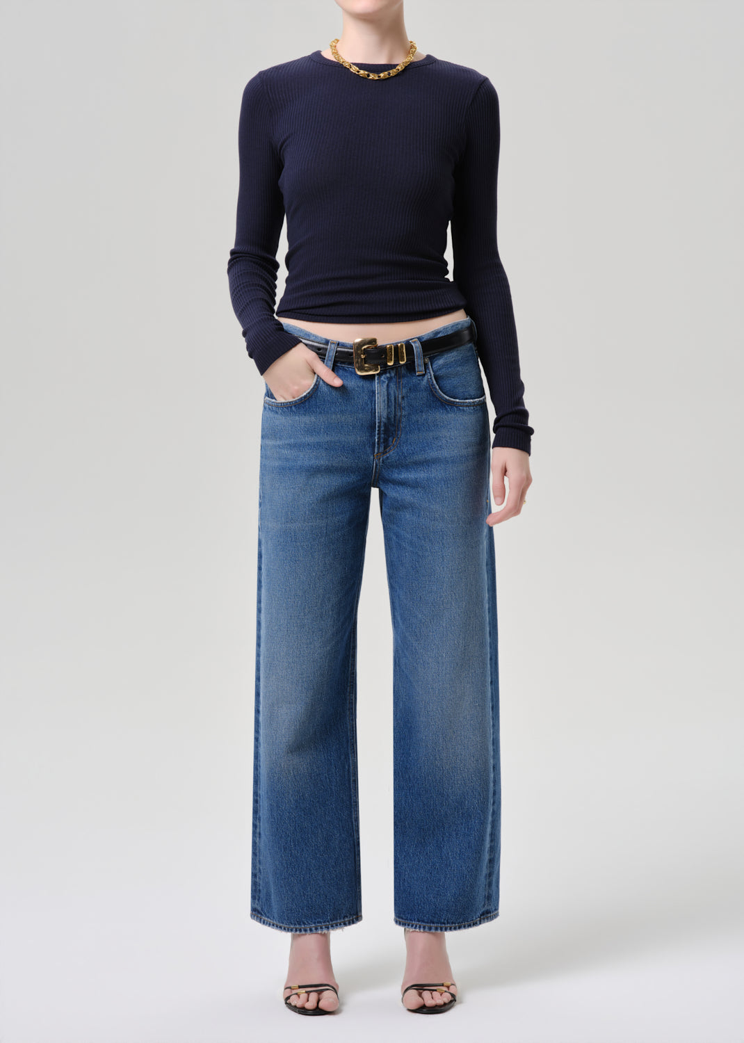 A model wearing a navy blue long-sleeve top, blue wide-leg denim jeans with a gold-tone belt, and black sandals, standing against a grey background.