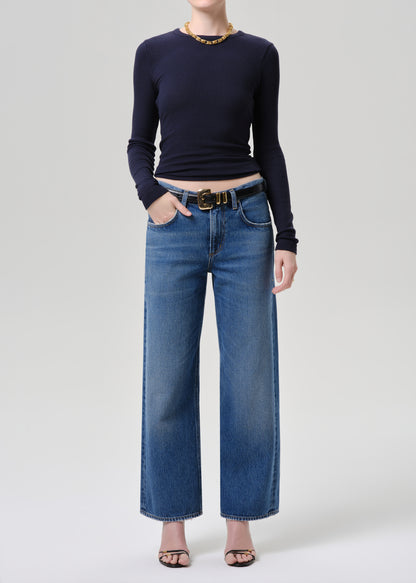 A model wearing a navy blue long-sleeve top, blue wide-leg denim jeans with a gold-tone belt, and black sandals, standing against a grey background.
