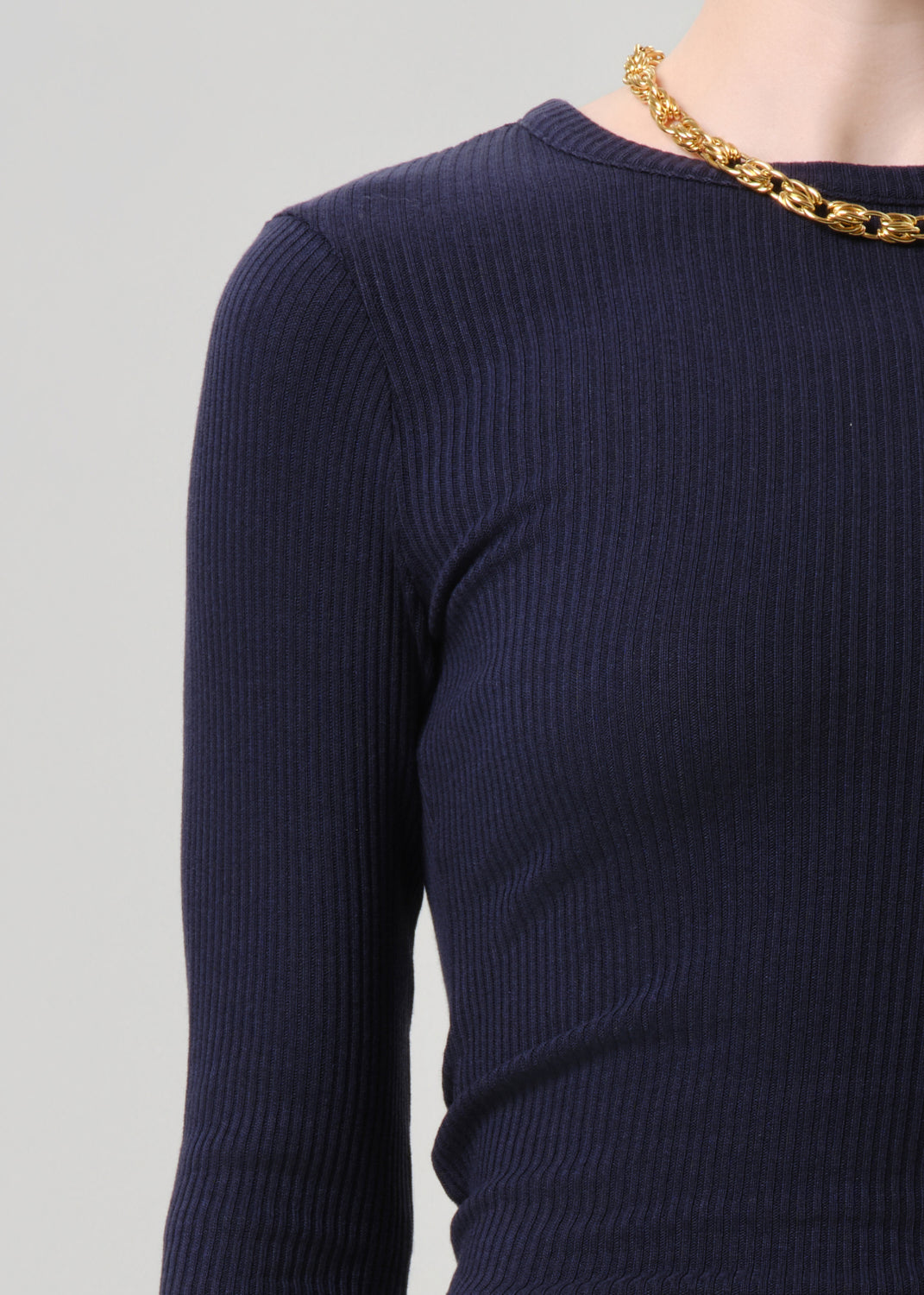 Close-up of a person wearing a dark navy ribbed long-sleeve top with a chunky gold chain necklace, against a grey background.