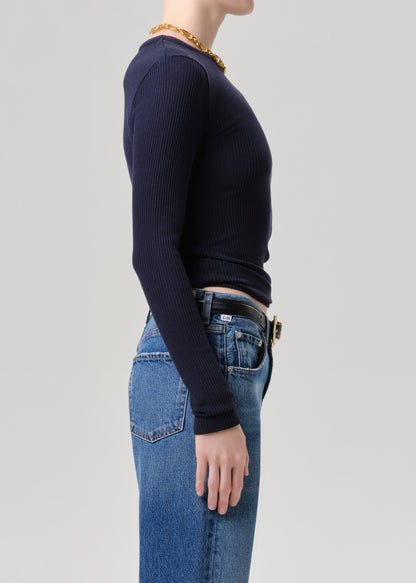 A side-profile view of a model wearing a dark navy ribbed long-sleeve top tucked into blue jeans with a black belt, against a grey background.