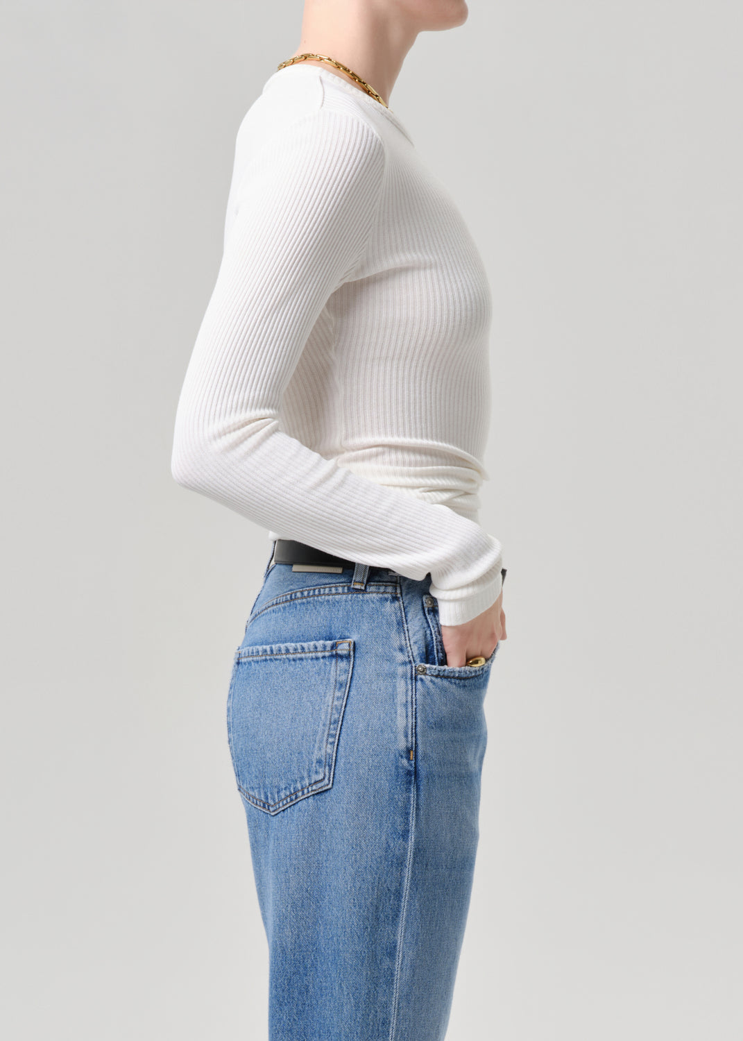 A side-profile view of a model wearing a white ribbed long-sleeve top and light blue jeans against a grey background.