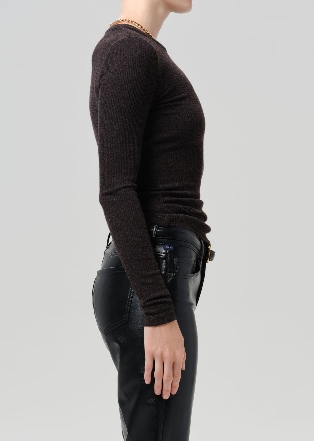 A side-profile view of a model wearing a dark brown long-sleeve knit sweater and black leather pants against a grey background.