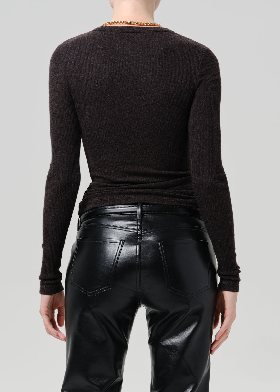 Rear view of a model wearing a dark brown long-sleeve knit top and black faux-leather pants against a grey background.