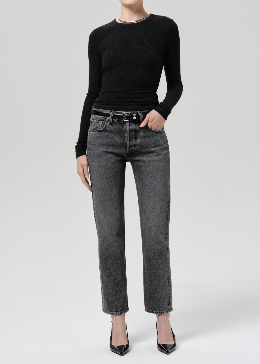A model wearing a black long-sleeve crewneck top, dark grey straight-leg denim jeans with a black leather belt, and black pointed-toe heels, photographed against a grey background.