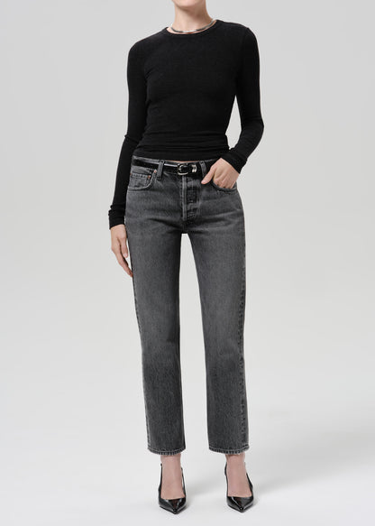 A model wearing a black long-sleeve crewneck top, dark grey straight-leg denim jeans with a black leather belt, and black pointed-toe heels, photographed against a grey background.