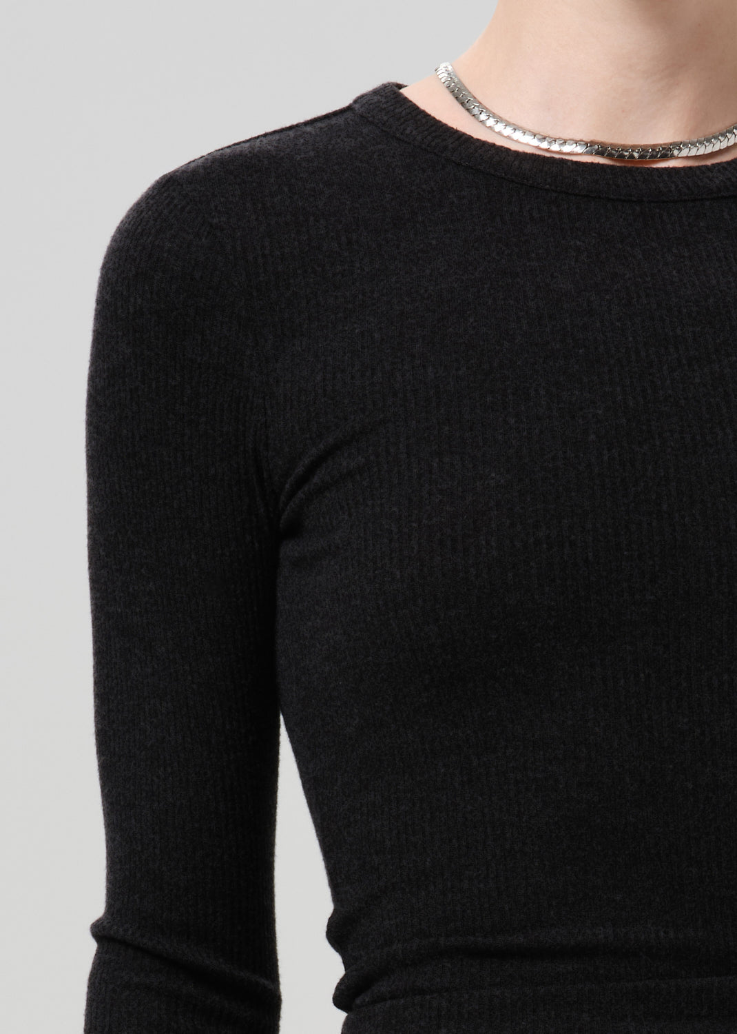 Close-up of a person wearing a black ribbed long-sleeve top with a silver chain necklace, against a grey background.