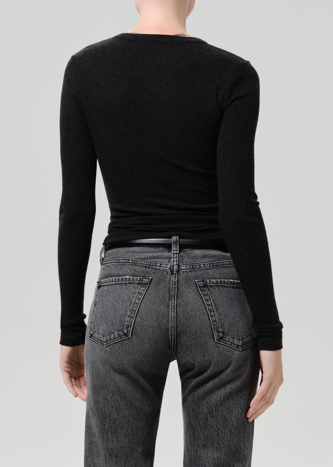 Rear view of a model wearing a black long-sleeve crewneck top tucked into dark grey washed jeans against a grey background.