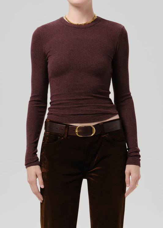 A model wearing a ribbed long-sleeve crewneck sweater in a deep burgundy brown, paired with dark brown velvet trousers and a black belt with a gold buckle, against a solid grey background.