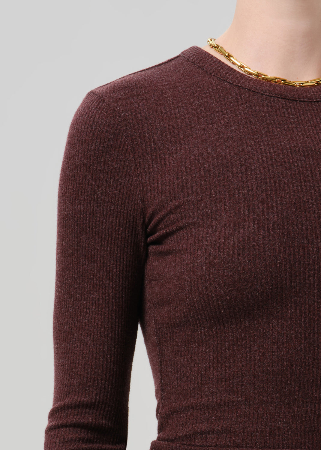Close-up of a person wearing a ribbed long-sleeve crew-neck sweater in dark burgundy, with a chunky gold chain necklace visible at the neckline, against a grey background.