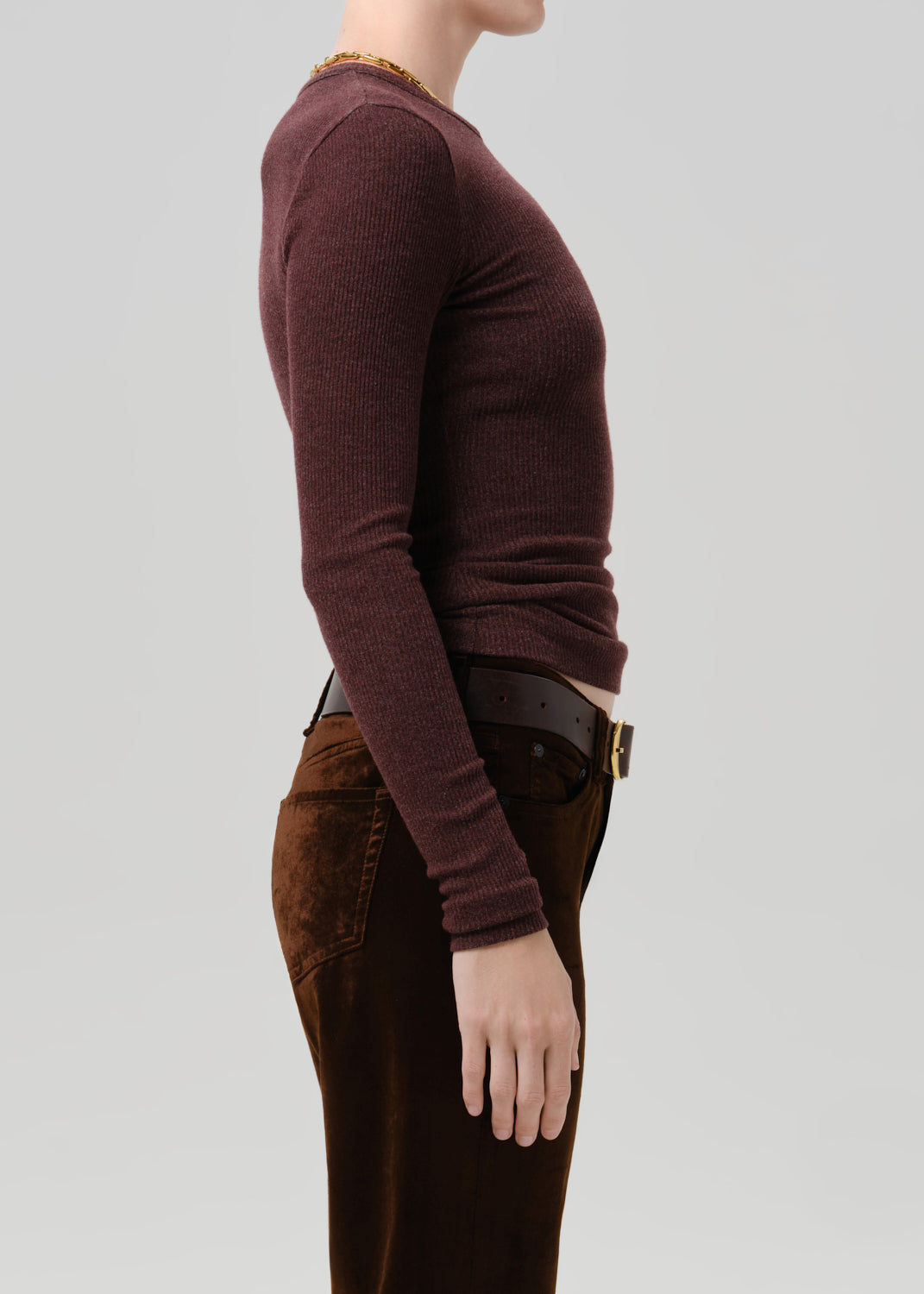 Side profile of a model wearing a ribbed long-sleeve crewneck sweater in a deep plum-burgundy color, paired with dark brown pants and a black belt, against a solid grey background.