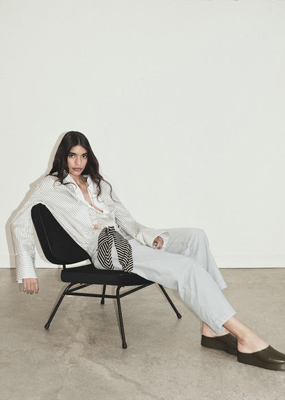 Woman sitting on a chair wearing a white shirt and light gray pants in a minimalistic setting.