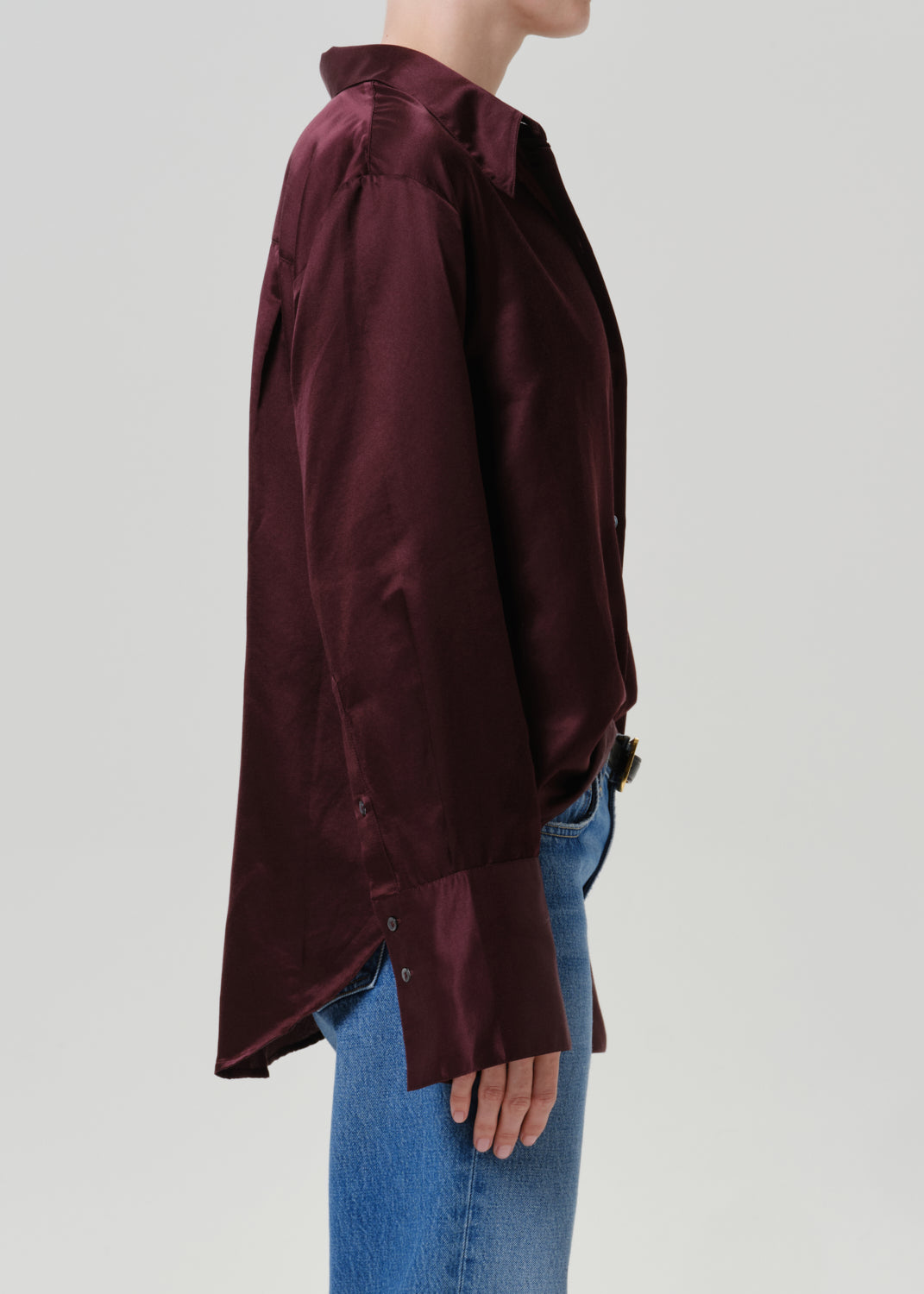 Woman wearing a burgundy satin button-up shirt with medium blue straight leg jeans viewed from the side on a grey background.