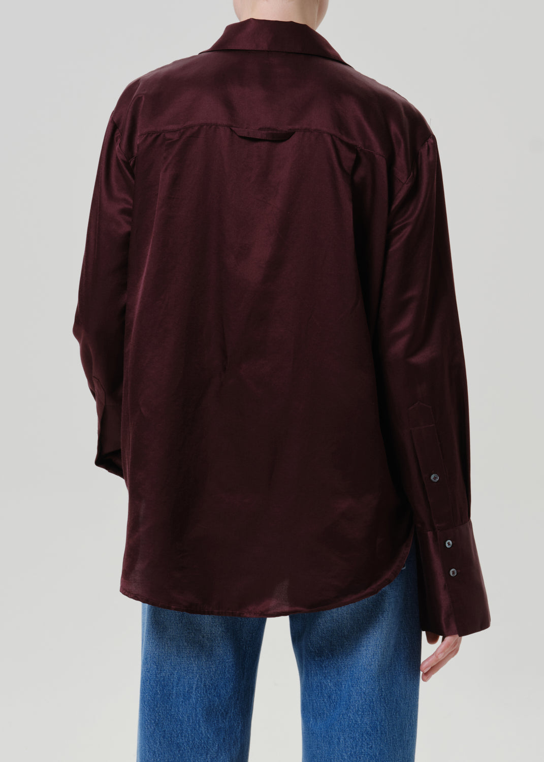 Woman wearing a burgundy satin button-up shirt with medium blue straight leg jeans viewed from the back on a grey background.