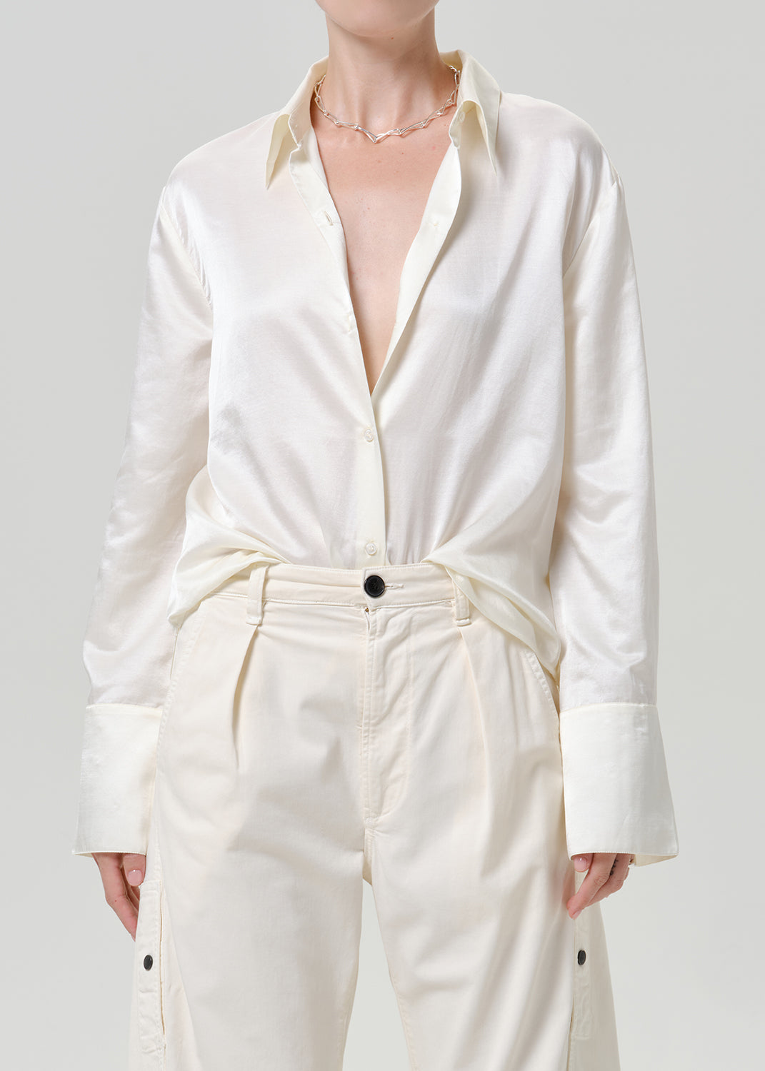 Woman wearing a white satin button-up shirt with white wide leg pants on a grey background.