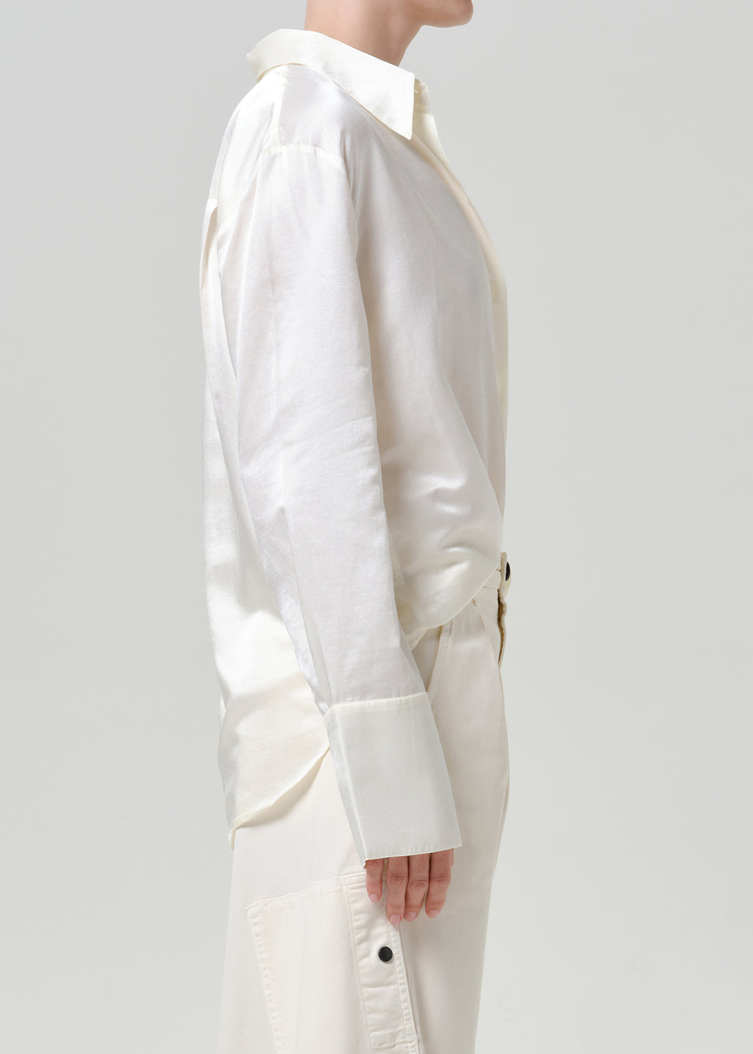 Woman wearing a white satin button-up shirt with white wide leg pants viewed from the side on a grey background.