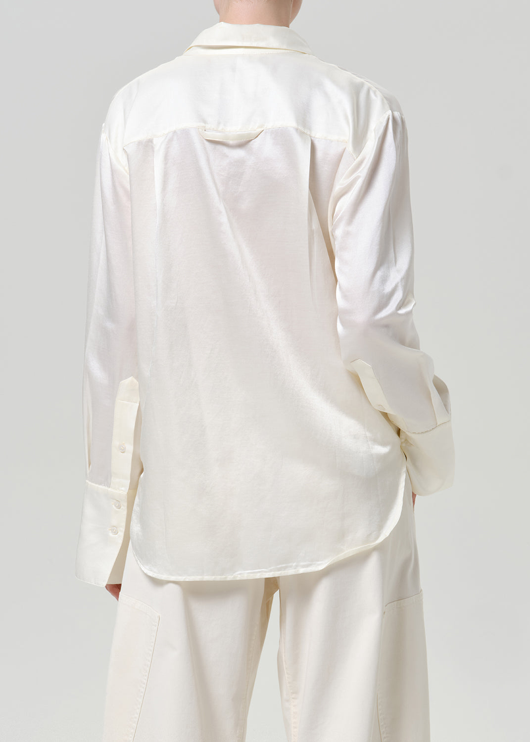 Woman wearing a white satin button-up shirt with white wide leg pants viewed from the back on a grey background.