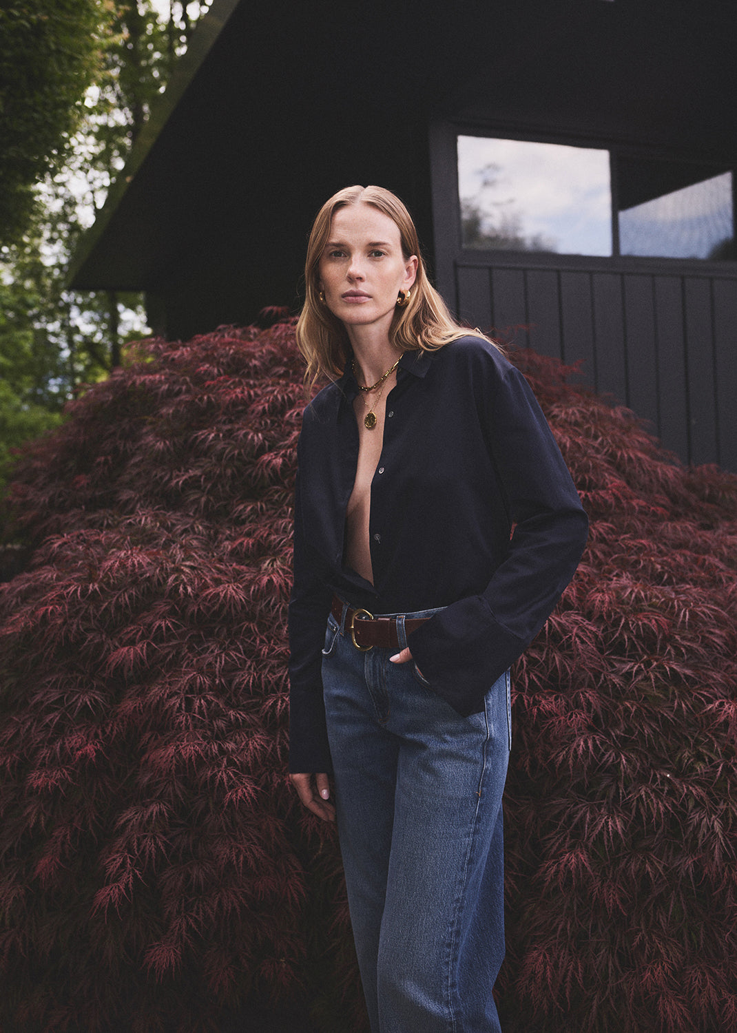 Woman standing outdoors with a dark building and greenery in the background