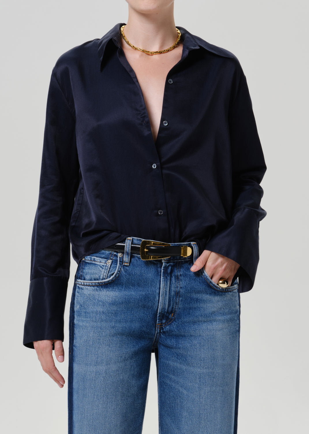 Woman wearing a dark navy satin button-up shirt with medium blue straight leg jeans on a grey background.