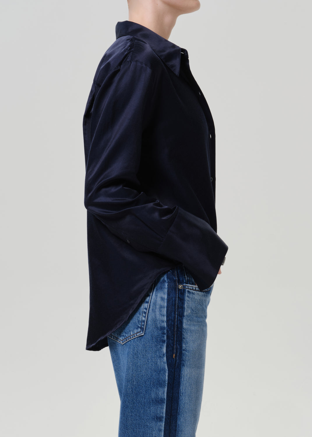 Woman wearing a dark navy satin button-up shirt with medium blue straight leg jeans viewed from the side on a grey background.