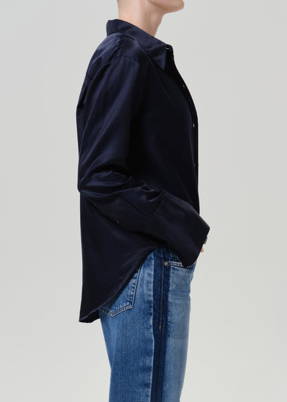 Woman wearing a dark navy satin button-up shirt with medium blue straight leg jeans viewed from the side on a grey background.