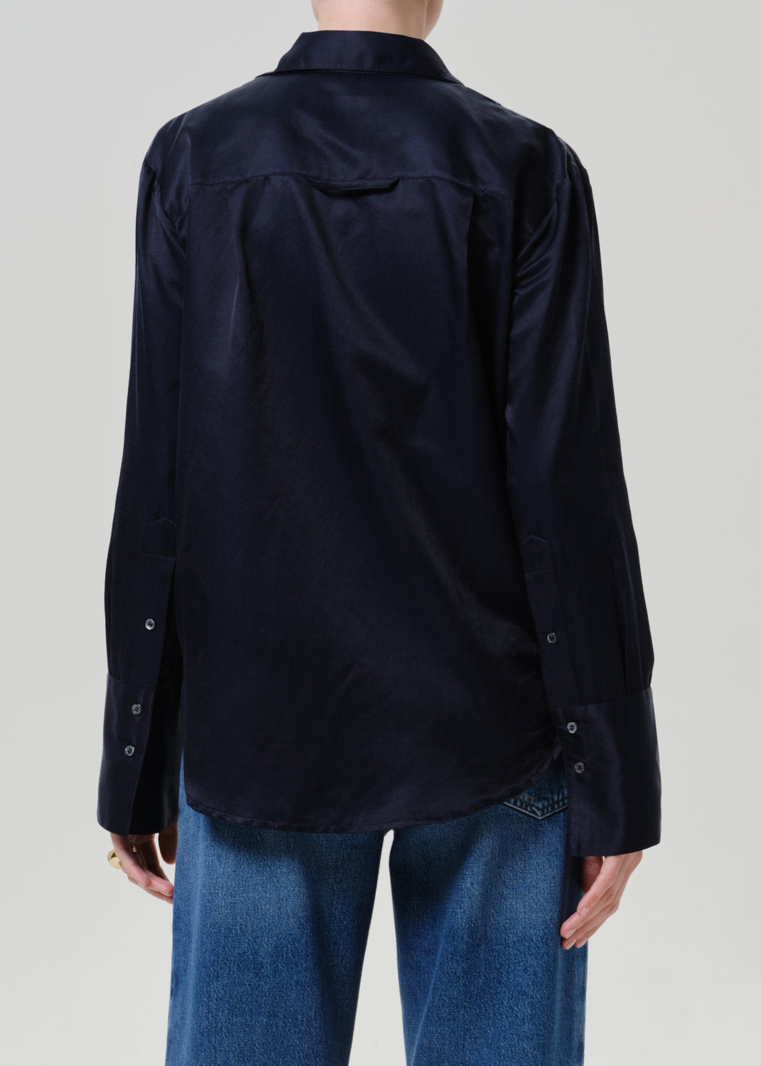 Woman wearing a dark navy satin button-up shirt with medium blue straight leg jeans viewed from the back on a grey background.