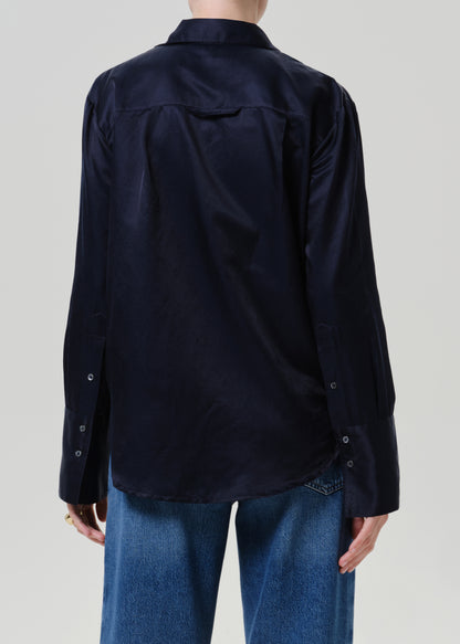 Woman wearing a dark navy satin button-up shirt with medium blue straight leg jeans viewed from the back on a grey background.