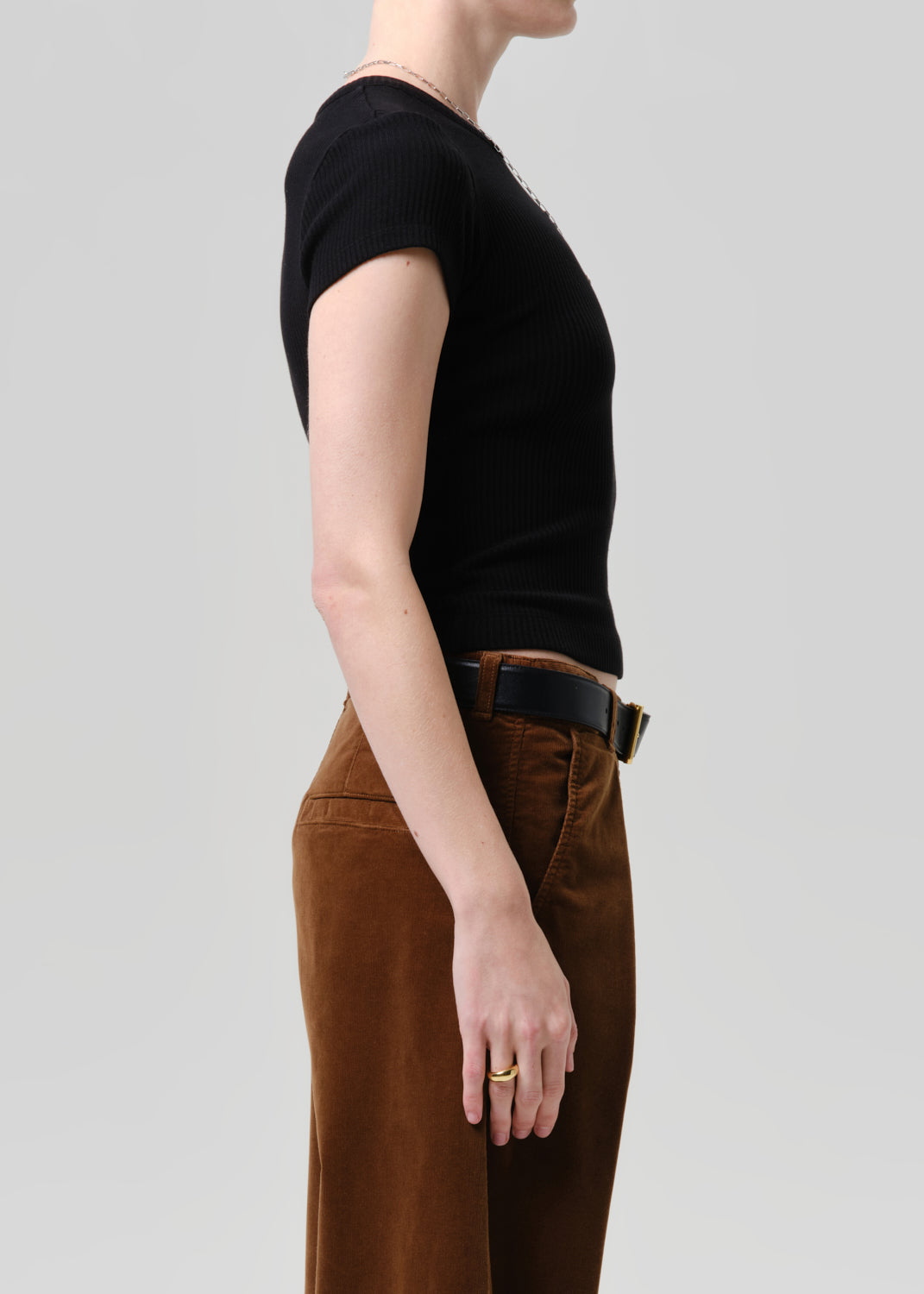 Side profile of a model wearing a black short-sleeve ribbed knit top and brown wide-leg trousers with a black belt, against a grey background.