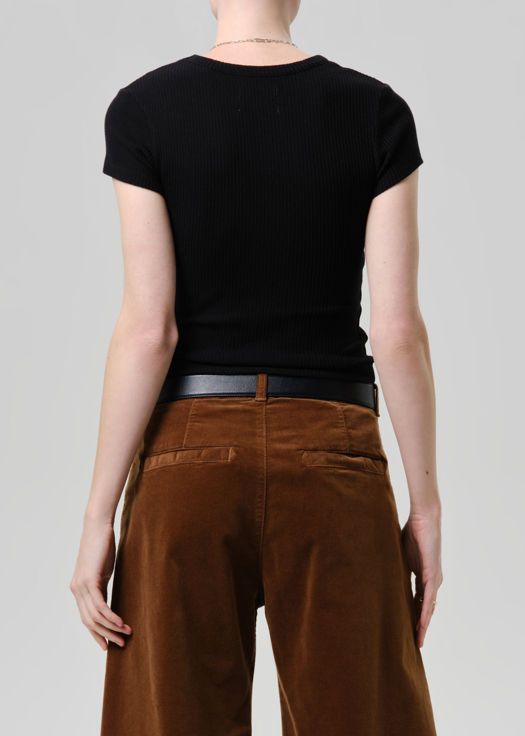 Rear view of a model wearing a black short-sleeve ribbed t-shirt and brown wide-leg trousers with a black belt, against a grey background.