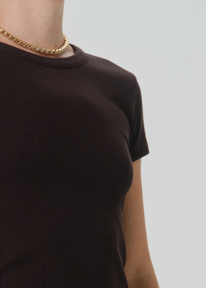 Close-up of a person wearing a dark brown ribbed short-sleeve crewneck top and a gold chain necklace against a grey background.