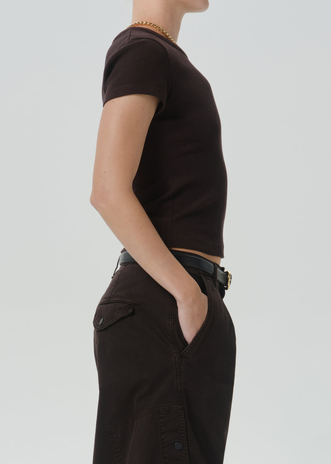 Side profile of a model wearing a dark brown short-sleeve crop top and dark brown wide-leg trousers with a black belt, set against a grey background.